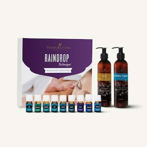 Op de afbeelding: Een Young Living Raindrop Technique-set met een paarse doos, 10 kleine flesjes etherische oliën en twee grotere flesjes massageolie. De doos heeft de tekst "Raindrop Technique" en "Rejuvenate the mind - Balance the body". De grotere flesjes zijn gelabeld met "V-6" en "Ortho Ease".