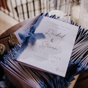 以下が含まれることがあります： 青いリボンをあしらった白い結婚式プログラムの束。一番上のプログラムには、「Rachel & Steven の結婚式へようこそ」というテキストと、「2024 年 4 月 27 日」の日付、「Cortlandt, NY」の場所が記載されています。