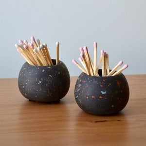 May include: Two black ceramic matchstick holders with speckled white and orange accents. Each holder is filled with wooden matches with pink tips.
