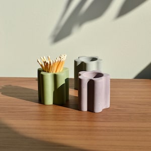 May include: Three concrete flower-shaped candle holders, one green and two purple, on a wooden surface. One holder has matches inside.