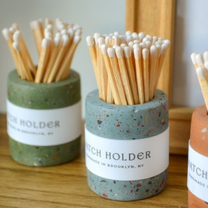 May include: Three terrazzo match holders with white tipped matches. The holders are green, blue, and orange. The labels on the holders read "Match Holder" and "Handmade in Brooklyn, NY".