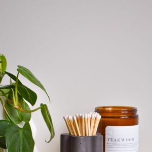 May include: A gray concrete matchstick holder with a star-shaped design, filled with wooden matches. A brown glass candle jar with a white label that reads "TEAKWOOD" is next to the holder. The items are on a table with a stack of magazines.