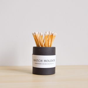 May include: A black match holder with a white label that reads "MATCH HOLDER HANDMADE IN BROOKLYN, NY." The holder is filled with wooden matches with white tips. The match holder is on a light brown wooden surface.