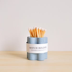 May include: A light blue concrete match holder with a white label that reads "Match Holder Handmade in Brooklyn, NY". The holder is filled with matches.