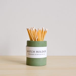 May include: A green concrete match holder with a white label that reads "Match Holder Handmade in Brooklyn, NY". The holder is filled with wooden matches.