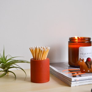 May include: A terracotta matchstick holder with a bundle of matches inside, a lit amber glass candle with a teakwood label, and a stack of magazines on a light wood surface.