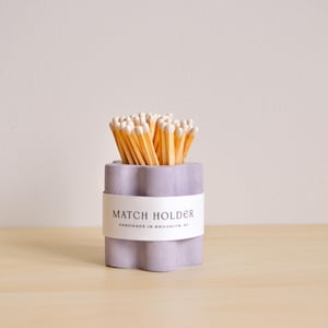 May include: A light purple concrete match holder with a white label that reads "MATCH HOLDER HANDMADE IN BROOKLYN, NY". The holder is filled with wooden matches.