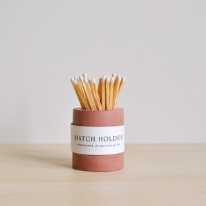 May include: A terracotta colored concrete match holder with a white label that reads "MATCH HOLDER HANDMADE IN BROOKLYN, NY". The holder is filled with wooden matches with white tips.