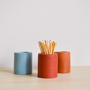 May include: Three concrete matchstick holders, one with matches inside. The holders are in blue, orange, and brown. The matches are light brown with white tips.