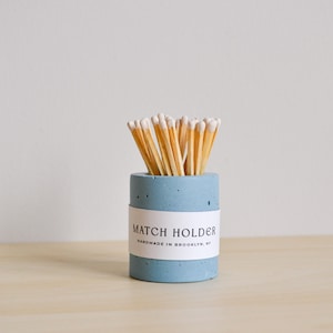May include: A light blue concrete match holder with a white label that reads "MATCH HOLDER HANDMADE IN BROOKLYN, NY". The holder is filled with wooden matches with white tips.