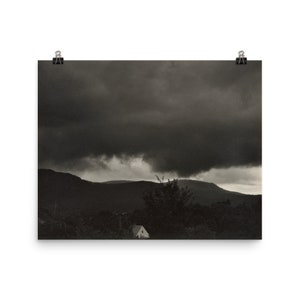 May include: A black and white photograph of a house in the distance with a dark, stormy sky above. The house is surrounded by trees and hills.
