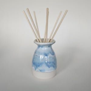 May include: A ceramic essential oil diffuser with a light blue and white glaze. The vase-shaped container holds several light-coloured wooden reeds, designed to release fragrance. The base is a solid white colour.