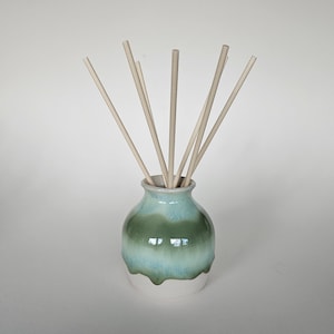 May include: A ceramic essential oil diffuser with a green and blue glaze gradient and a white base. The vase holds several light-coloured wooden reeds, designed to release fragrance. Home decor item.