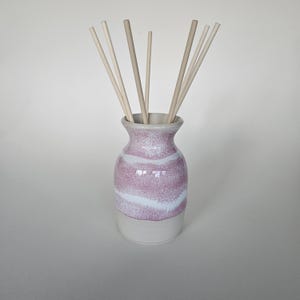 May include: A ceramic essential oil diffuser with a pink and white speckled design. The vase-shaped container has a light grey base and holds several light-coloured wooden diffuser sticks.