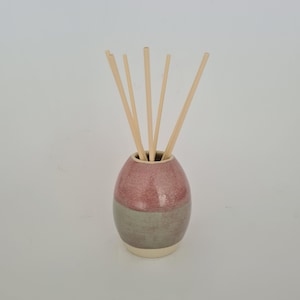 May include: A two-tone ceramic essential oil diffuser. The upper section is pink, the lower section is grey-green. Several light-coloured wooden reeds are inserted, designed to release fragrance. The base is a light cream colour. The diffuser is approximately 12 cm tall.