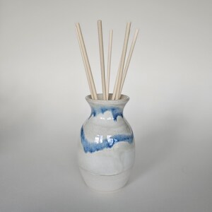 May include: A ceramic essential oil diffuser with a white and blue glazed finish. The vase holds several light-coloured wooden reeds. The design features a rounded base and a flared opening, suitable for home fragrance.