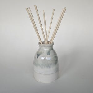 May include: A ceramic reed diffuser in a light grey and white glaze. The vase contains several light-coloured wooden reeds. The base of the vase is a solid white colour. The diffuser is designed for home fragrance.