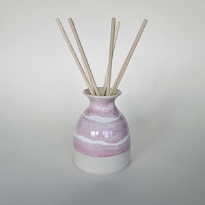 May include: A ceramic essential oil diffuser with a pink and white striped design. The vase has a white base and holds several wooden reeds. This decorative item is designed for home fragrance.