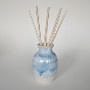 May include: A ceramic essential oil diffuser with a light blue and white glaze. The vase-shaped container holds several light-coloured wooden reeds. The bottom of the vase is white. The diffuser is designed for home fragrance.