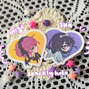 May include: Two heart-shaped stickers with anime-style illustrations. One features a pink-haired character on a yellow background, the other a dark-haired character on a purple background. The text "mizi" and "sua" are visible. The stickers are surrounded by star-shaped origami.