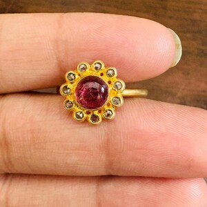 Puede incluir: Un anillo de oro con una piedra preciosa central, redonda y de color rojo intenso, rodeada de piedras preciosas más pequeñas y claras. El diseño del anillo se asemeja a una flor, con las piedras engastadas en una banda de oro.
