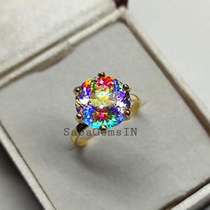 May include: A gold ring featuring a large, round, multi-coloured gemstone. The gemstone displays a rainbow of colours, including red, orange, yellow, green, blue, and purple. The ring is set in a gold band with a simple design. The ring is presented in a white box with the text "SabaGems IN".