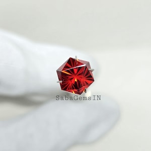 Puede incluir: Una gema roja facetada con un diseño geométrico. La gema está montada sobre un engaste plateado. El texto "SabaGems IN" es visible debajo.