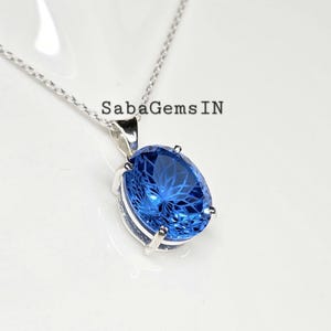 May include: A silver necklace with a large, oval-shaped, bright blue gemstone pendant. The gemstone is faceted, catching the light. The text "SabaGems IN" is visible on the image.