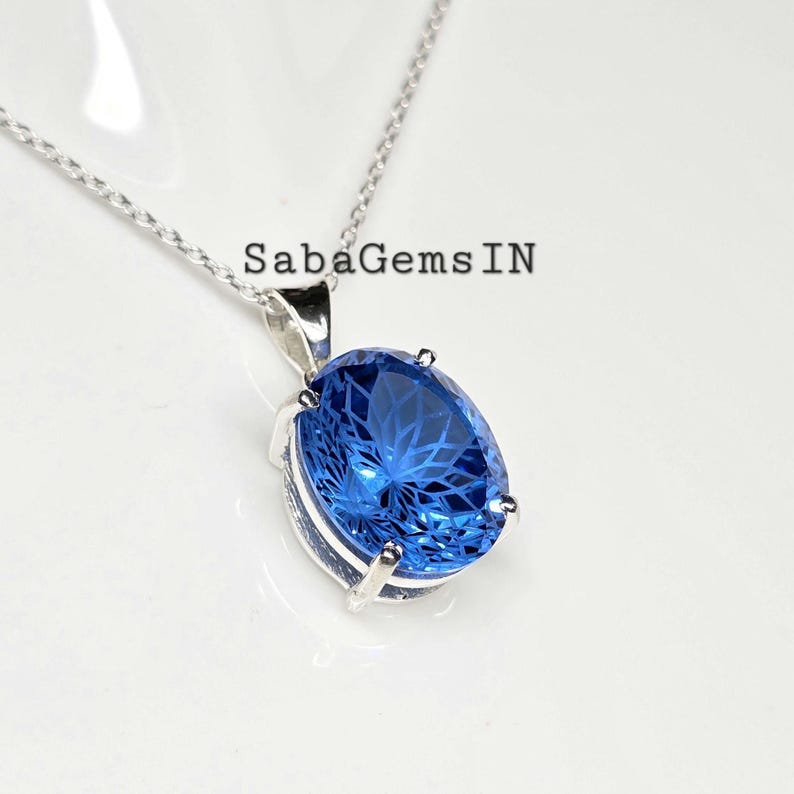May include: A silver necklace with a large, oval-shaped, bright blue gemstone pendant. The gemstone is faceted, catching the light. The text "SabaGems IN" is visible on the image.