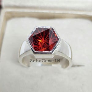 May include: A silver ring with a red hexagonal gemstone. The gemstone has a starburst pattern and is set in a silver bezel. The ring is in a white box with the text "SabaGemsIN" on the bottom.