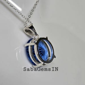 May include: A silver necklace with a pendant featuring a large, oval-shaped, faceted blue gemstone. The pendant is encased in a silver setting and hangs from a delicate chain. The text "925" is visible on the pendant's bail. The brand name "SabaGems IN" is at the bottom.
