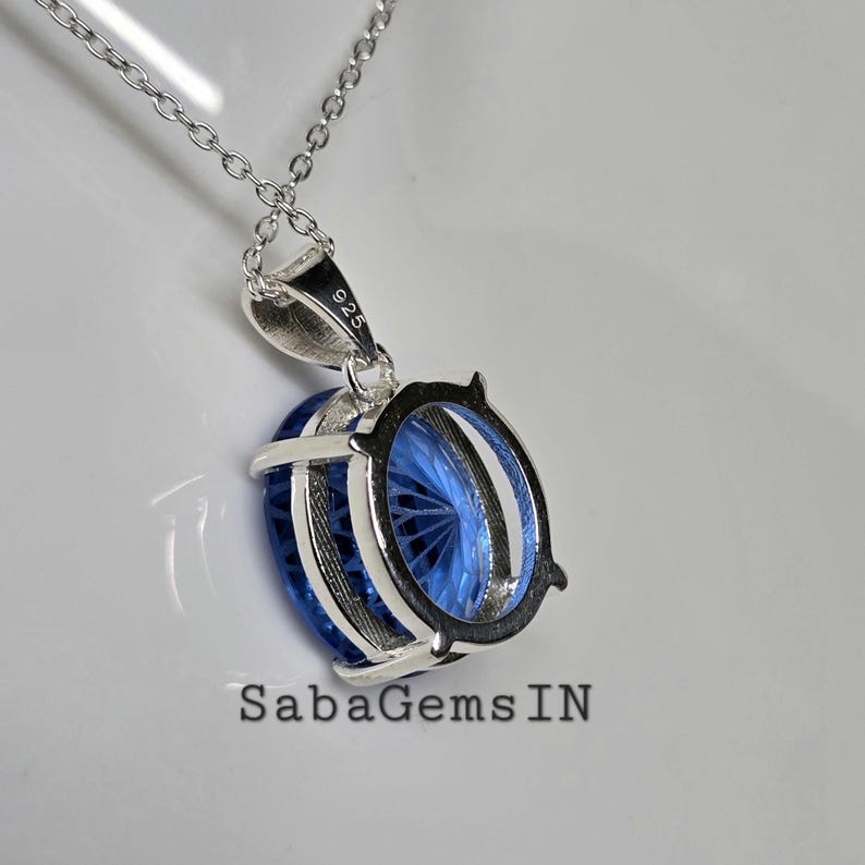 May include: A silver necklace with a pendant featuring a large, oval-shaped, faceted blue gemstone. The pendant is encased in a silver setting and hangs from a delicate chain. The text "925" is visible on the pendant's bail. The brand name "SabaGems IN" is at the bottom.