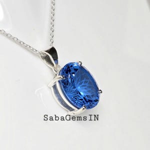 May include: A silver necklace with a large, oval, faceted blue gemstone pendant. The pendant is set in a silver bezel and hangs from a delicate silver chain. The text "SabaGemsIN" is visible.