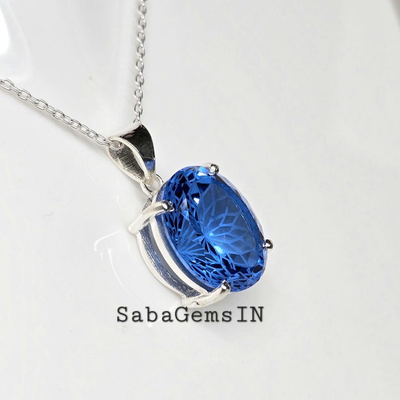 May include: A silver necklace with a large, oval, faceted blue gemstone pendant. The pendant is set in a silver bezel and hangs from a delicate silver chain. The text "SabaGemsIN" is visible.