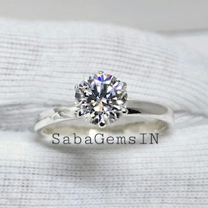 Könnte beinhalten: Ein silberner Ring mit einem großen, runden, klaren Diamanten im Zentrum. Der Ring liegt auf einem weißen Stoffhintergrund. Der Text "SabaGems IN" ist unterhalb des Rings sichtbar.
