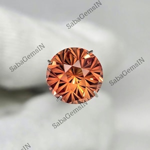 May include: A round, faceted gemstone with a warm orange hue. The gem is held in place by four silver prongs. The gem's facets create a starburst pattern, reflecting light. The background is a soft, neutral colour.