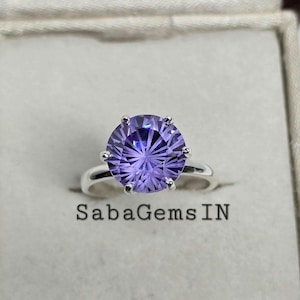 Op de afbeelding: Een zilveren ring met een grote, ronde, paarse edelsteen. De edelsteen heeft een stervormig patroon.