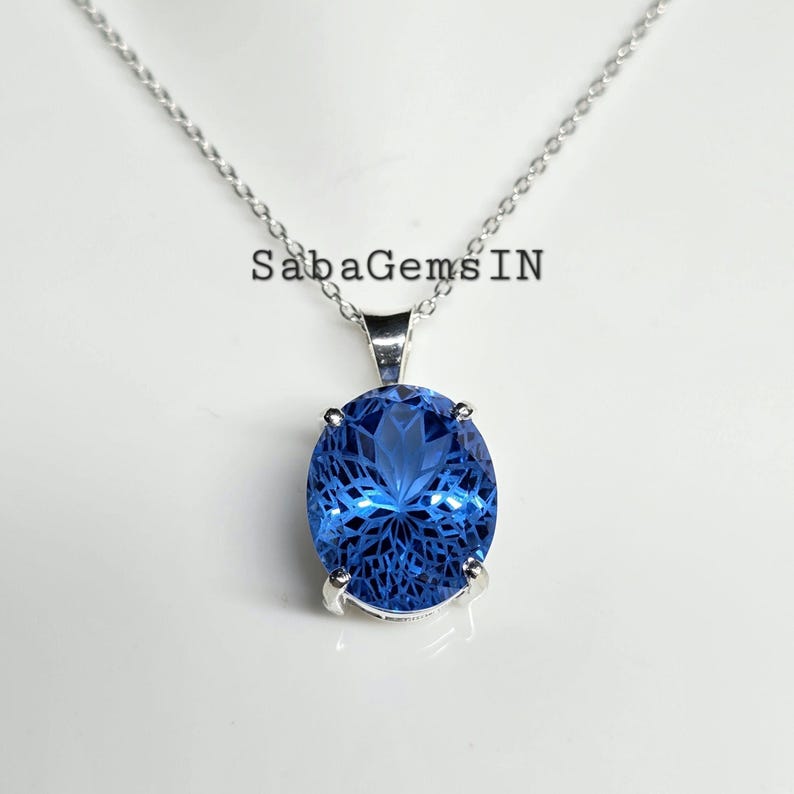 May include: A silver necklace with a large, oval, faceted blue gemstone pendant. The pendant is set in a silver bezel and hangs from a delicate silver chain. The text "SabaGemsIN" is visible.