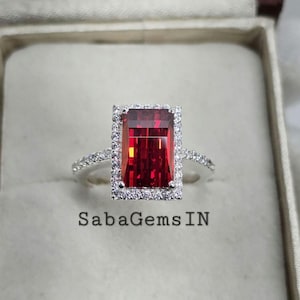 May include: A red gemstone ring with a square-shaped stone set in a halo of smaller diamonds. The ring is set in a silver band.