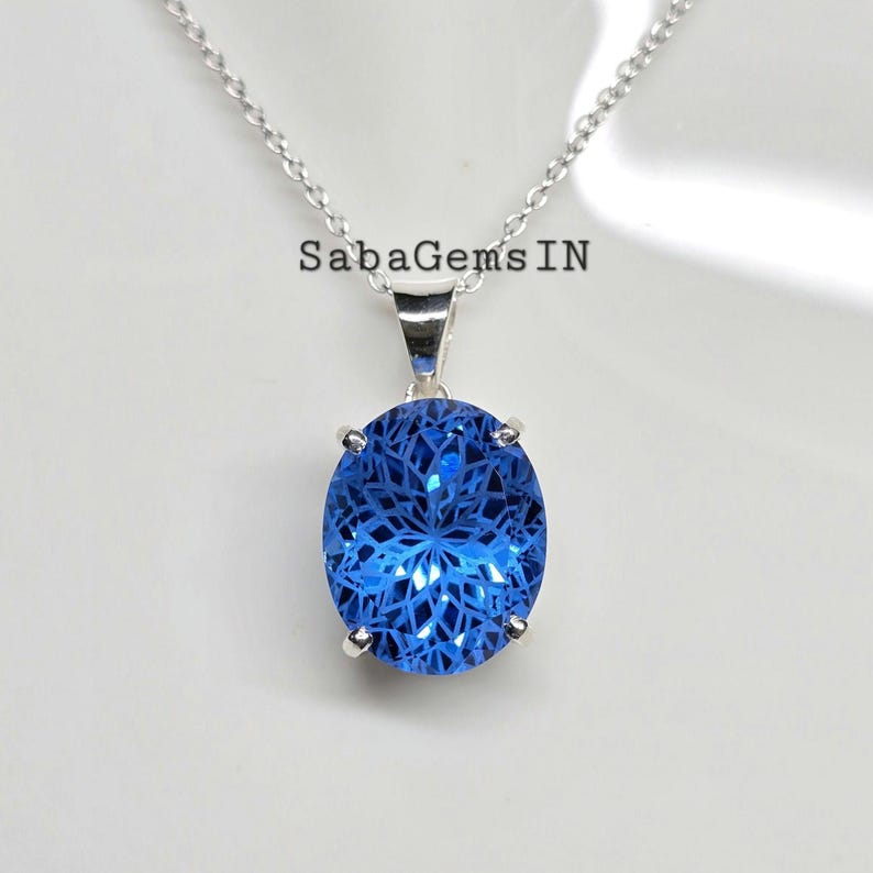 May include: A silver necklace with a large, oval, bright blue pendant. The pendant has a detailed, intricate pattern within the blue gemstone. The text "SabaGemsIN" is visible on the image.