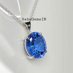May include: A silver necklace with a large, oval-shaped, bright blue gemstone pendant. The gemstone has a faceted design, and the pendant is set in a silver bezel. The necklace is displayed on a white surface.