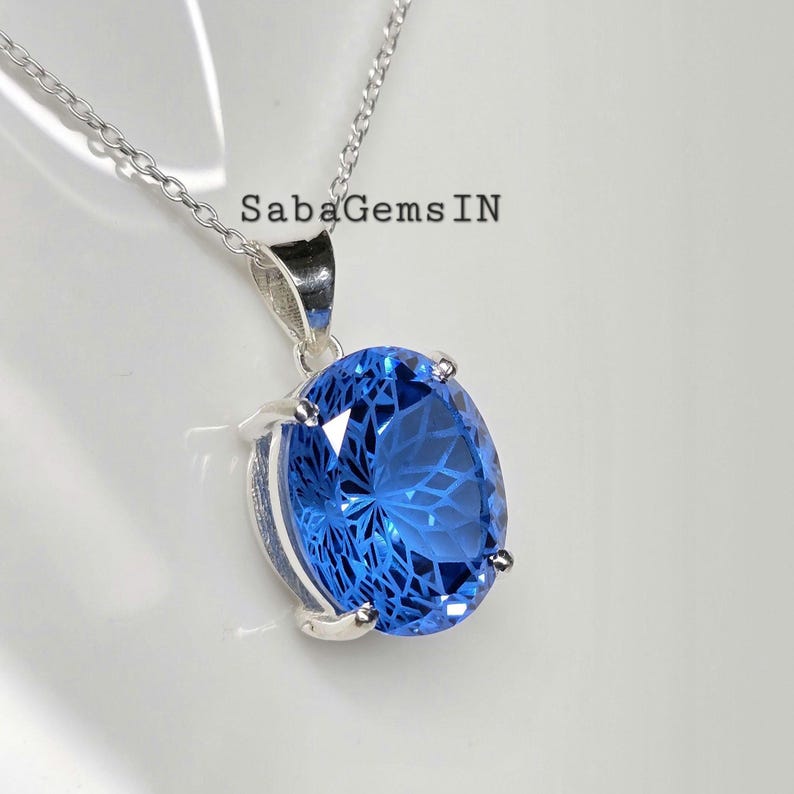 May include: A silver necklace with a large, oval-shaped, bright blue gemstone pendant. The gemstone has a faceted design, and the pendant is set in a silver bezel. The necklace is displayed on a white surface.