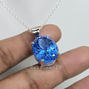 May include: A silver necklace featuring a large, oval-shaped blue gemstone pendant. The pendant has intricate internal facets, creating a sparkling effect. The chain is delicate, and the pendant is held by silver prongs. The text "SabaGems IN" is visible.