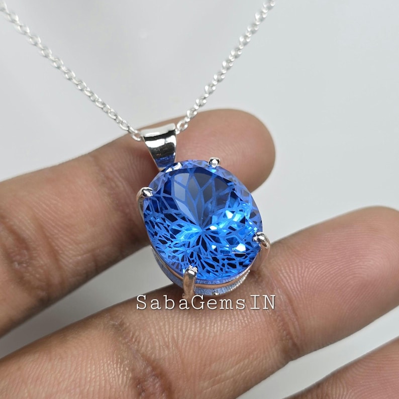 May include: A silver necklace featuring a large, oval-shaped blue gemstone pendant. The pendant has intricate internal facets, creating a sparkling effect. The chain is delicate, and the pendant is held by silver prongs. The text "SabaGems IN" is visible.