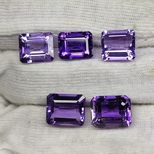 May include: Five rectangular, faceted amethyst gemstones. The gems are a deep purple color and have a polished, reflective surface. The stones are arranged on a white, textured fabric.