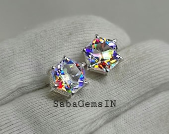 May include: Two star-shaped, silver-toned stud earrings with iridescent, multi-colored gemstones. The earrings are displayed on a white, textured surface. The text "SabaGemsIN" is visible.