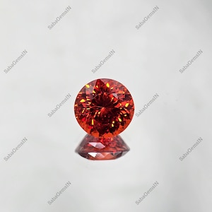 May include: A round, faceted, vibrant orange-red gemstone with a brilliant cut, displaying internal light reflections and a clear reflection of its bottom surface visible directly beneath it on a smooth, light-colored surface.