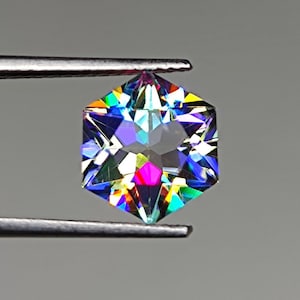 Könnte beinhalten: Ein sechseckiger, schillernder Edelstein mit einem Regenbogen aus Farben, darunter Blau, Grün, Gelb, Orange, Rot und Lila. Der Edelstein wird von einer Pinzette gehalten.