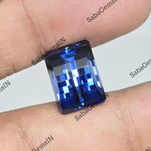 May include: A rectangular faceted gemstone with a deep royal blue colour and a high-gloss finish. The stone features a clear, geometric cut that reflects light, creating bright white highlights across its surface, showcasing a vibrant blue hue.