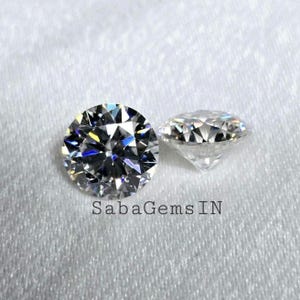 Könnte beinhalten: Zwei runde, klare, brillantgeschliffene Edelsteine auf einer weißen Oberfläche. Die Edelsteine sind in einer Zargenfassung gefasst. Der Text "SabaGems IN" ist unter den Edelsteinen sichtbar.
